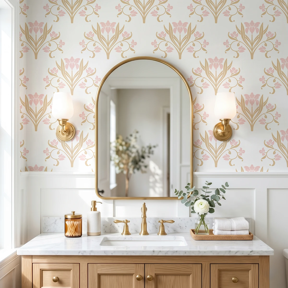 Liria, papel pintado floral mostaza suave y rosa empolvado, a escala grande en baño elegante con mueble de lavabo, espejo de arco y apliques de pared.