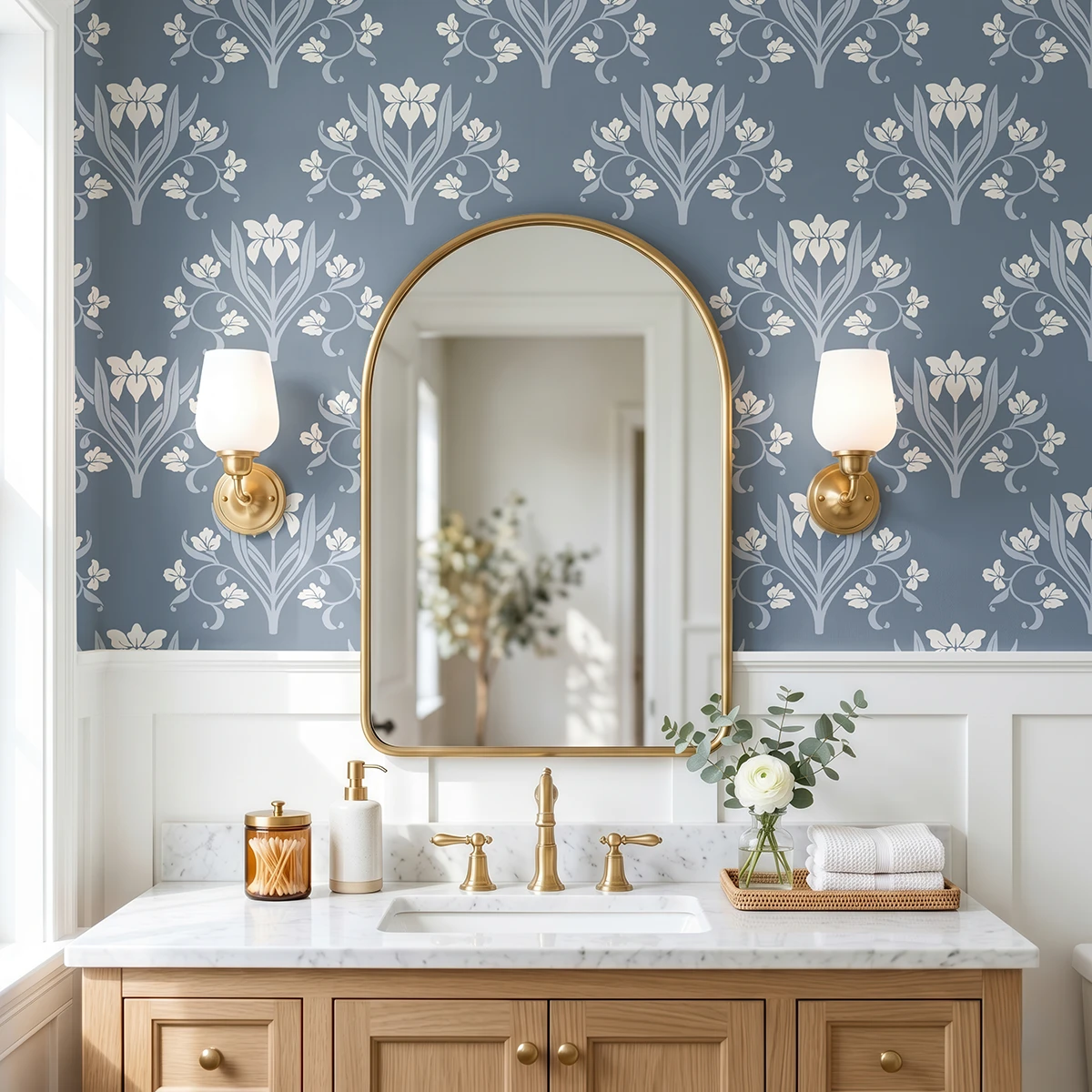 Liria, papel pintado floral azul noche y blanco roto, a escala grande en baño elegante con mueble de lavabo, espejo de arco y apliques de pared.