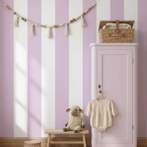 Godella, papel pintado de rayas lavanda y blanco, a escala maxi en habitación infantil con un armario alto de madera, del cual cuelga un mameluco beige en una percha. Delante de la pared hay un banco de madera con un peluche de oveja y una cesta de mimbre sobre el armario, todo decorado con una guirnalda de borlas de cuerda en la parte superior que aporta un toque artesanal y tierno..