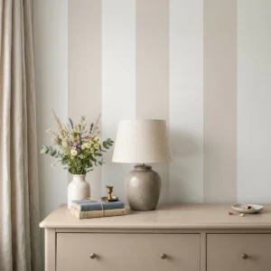 Modelo Della, papel pintado de rayas beige y blanco lino, a escala maxi en dormitorio con cómoda de madera clara, con una lámpara de sobremesa, algunos libros y un pequeño jarrón con flores.
