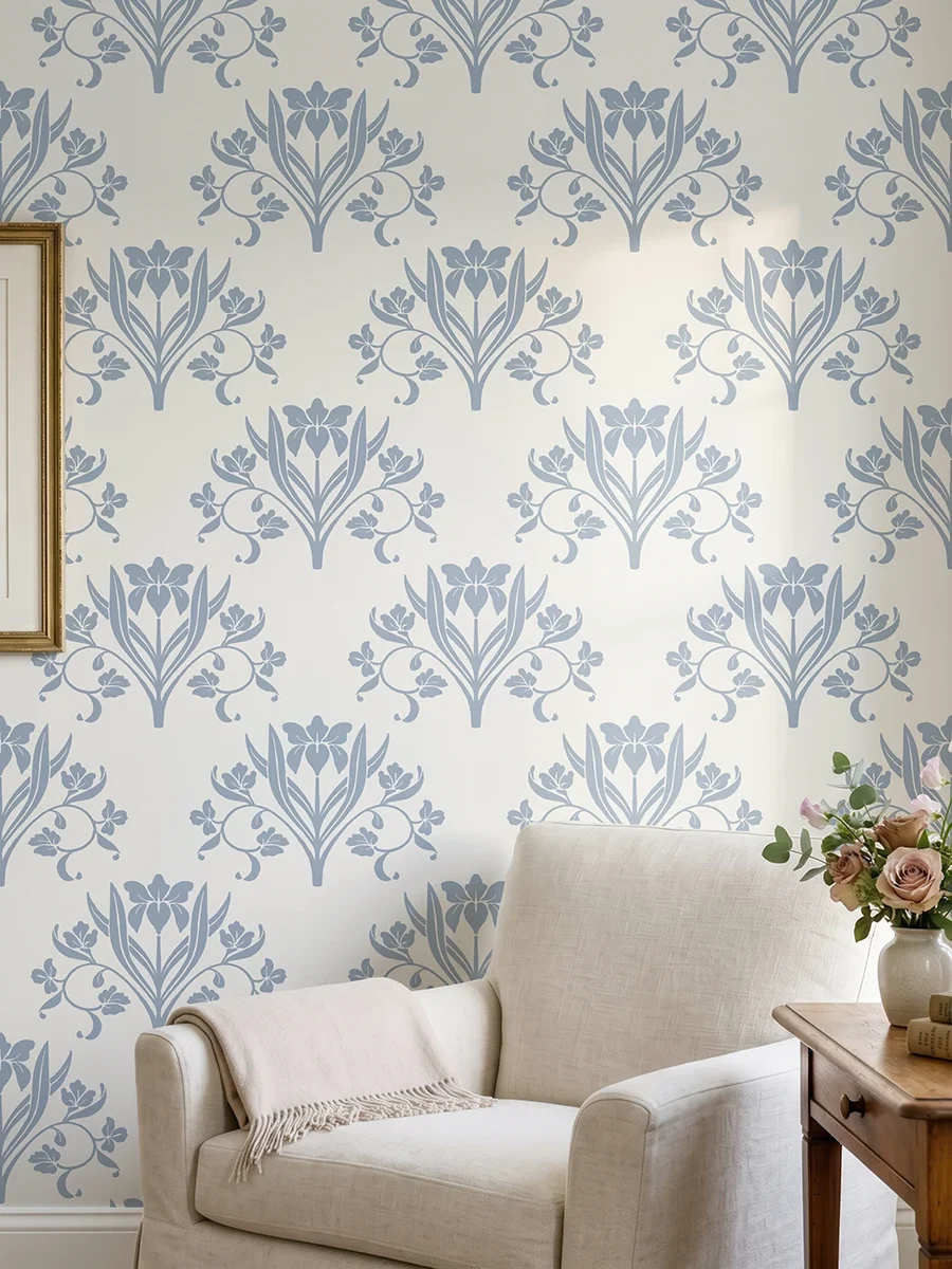 Liria, papel pintado floral azul empolvado y blanco lino, a escala grande en salón luminoso con butaca, decorado con cuadros y elementos vegetales.