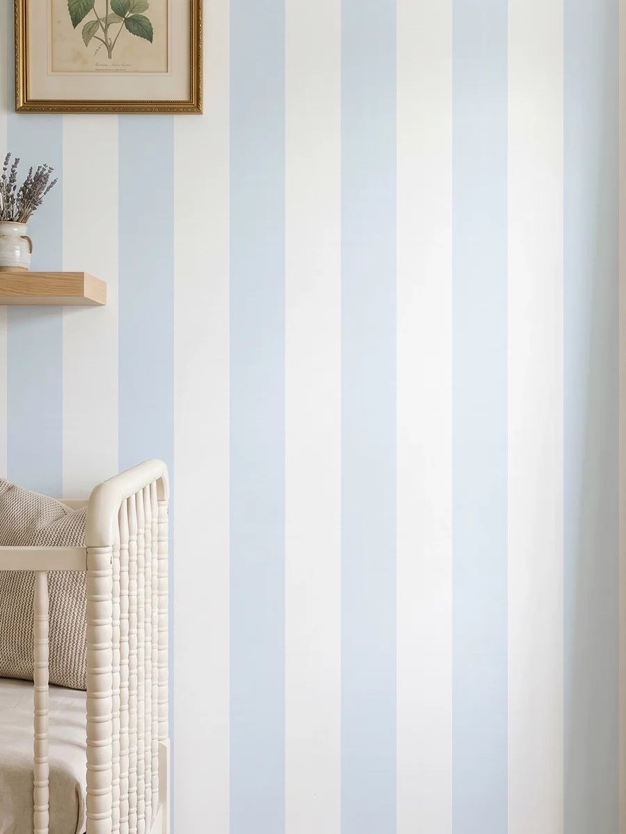 Modelo Della, papel pintado de rayas azul celeste y blanco, a escala grande en dormitorio infantil con cuna de bebé y pared decorada con una pequeña estantería con elementos decorativos.