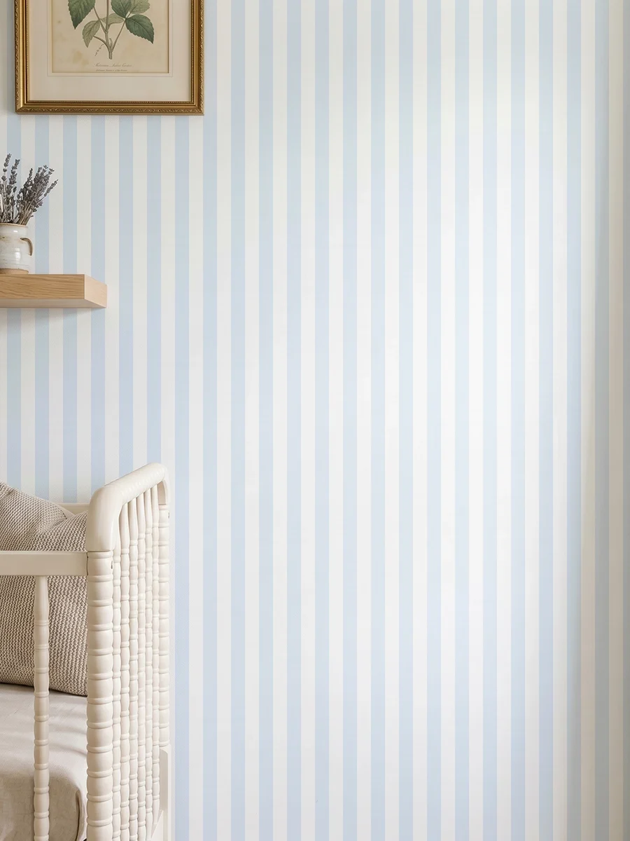 Modelo Della, papel pintado de rayas azul celeste y blanco, a escala pequeña en dormitorio infantil con cuna de bebé y pared decorada con una pequeña estantería con elementos decorativos.