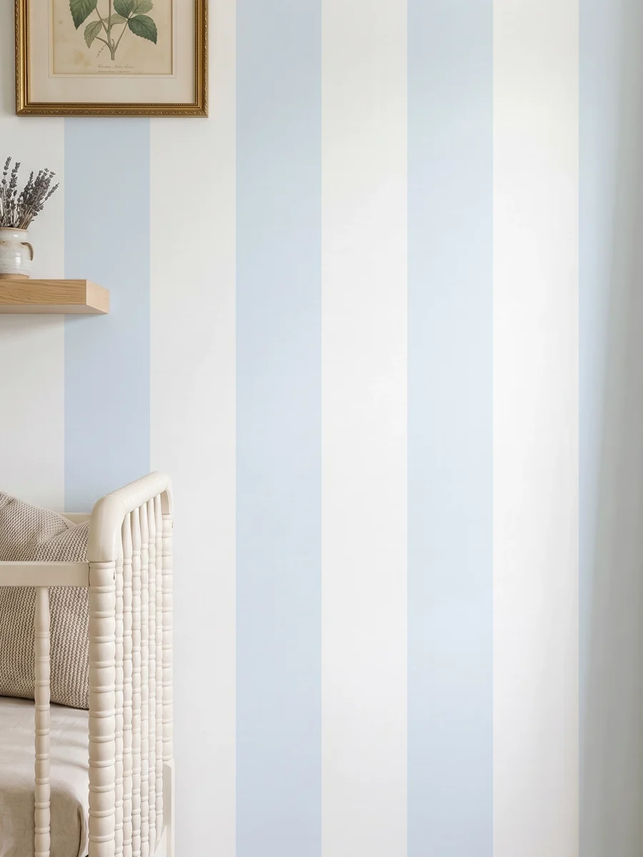 Modelo Della, papel pintado de rayas azul celeste y blanco, a escala maxi en dormitorio infantil con cuna de bebé y pared decorada con una pequeña estantería con elementos decorativos.