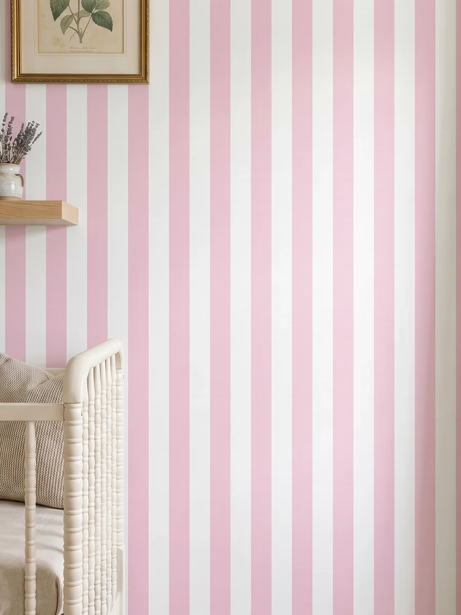 Modelo Della, papel pintado de rayas rosa rubor y blanco, a escala mediana en dormitorio infantil con cuna de bebé y pared decorada con una pequeña estantería con elementos decorativos.