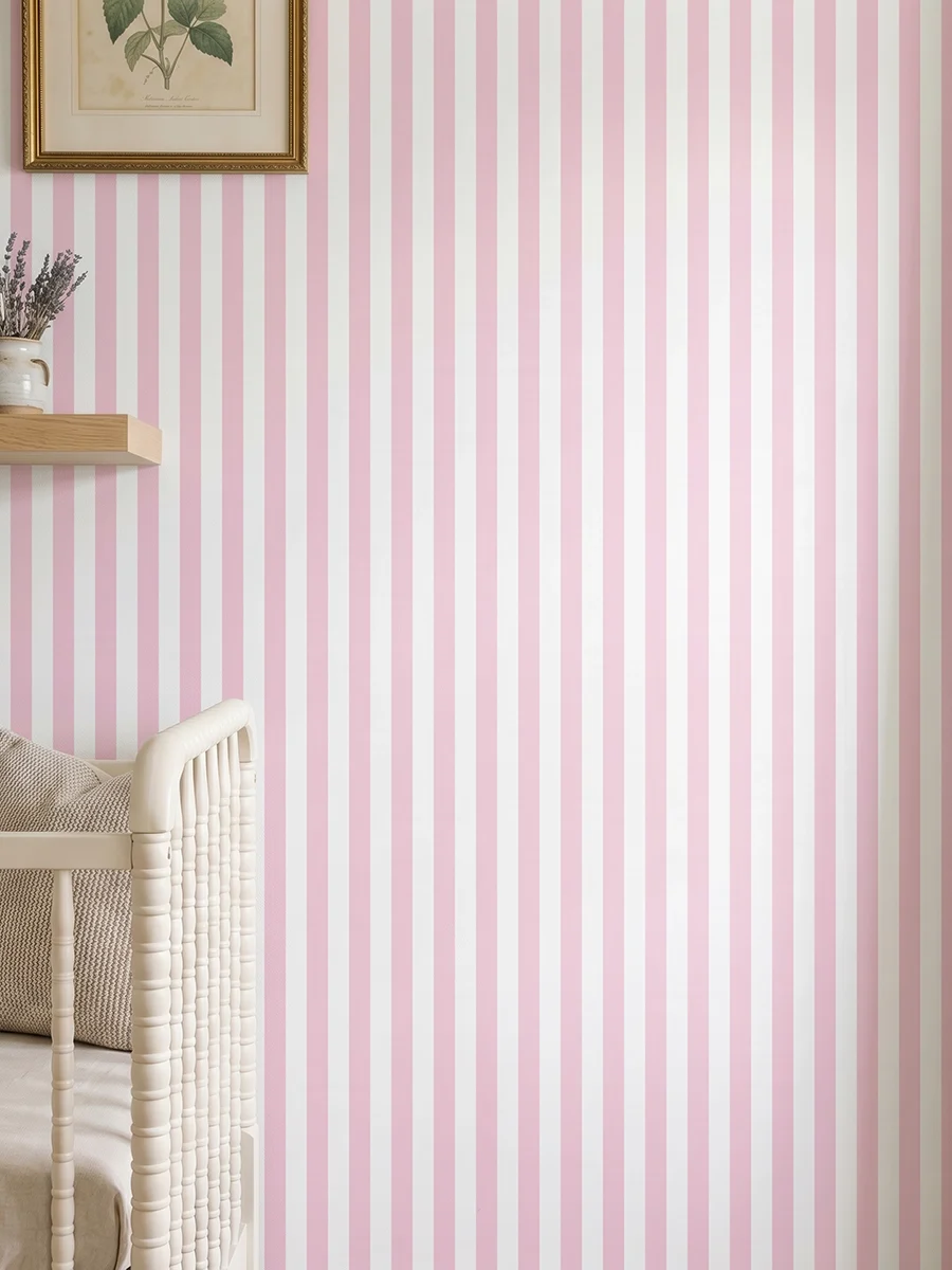 Modelo Della, papel pintado de rayas rosa rubor y blanco, a escala pequeña en dormitorio infantil con cuna de bebé y pared decorada con una pequeña estantería con elementos decorativos.