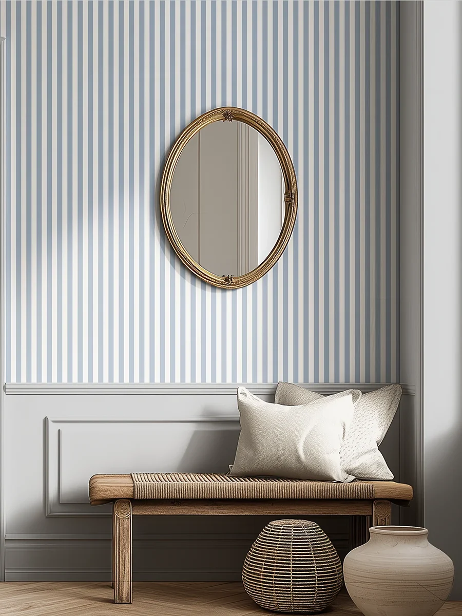 Modelo Della, papel pintado de rayas azul empolvado y blanco lino, a escala pequeña en pasillo o recibidor luminoso con un gran ventanal, espejo ovalado con marco dorado, un banco coqueto con cojines y elementos decorativos artesanales.