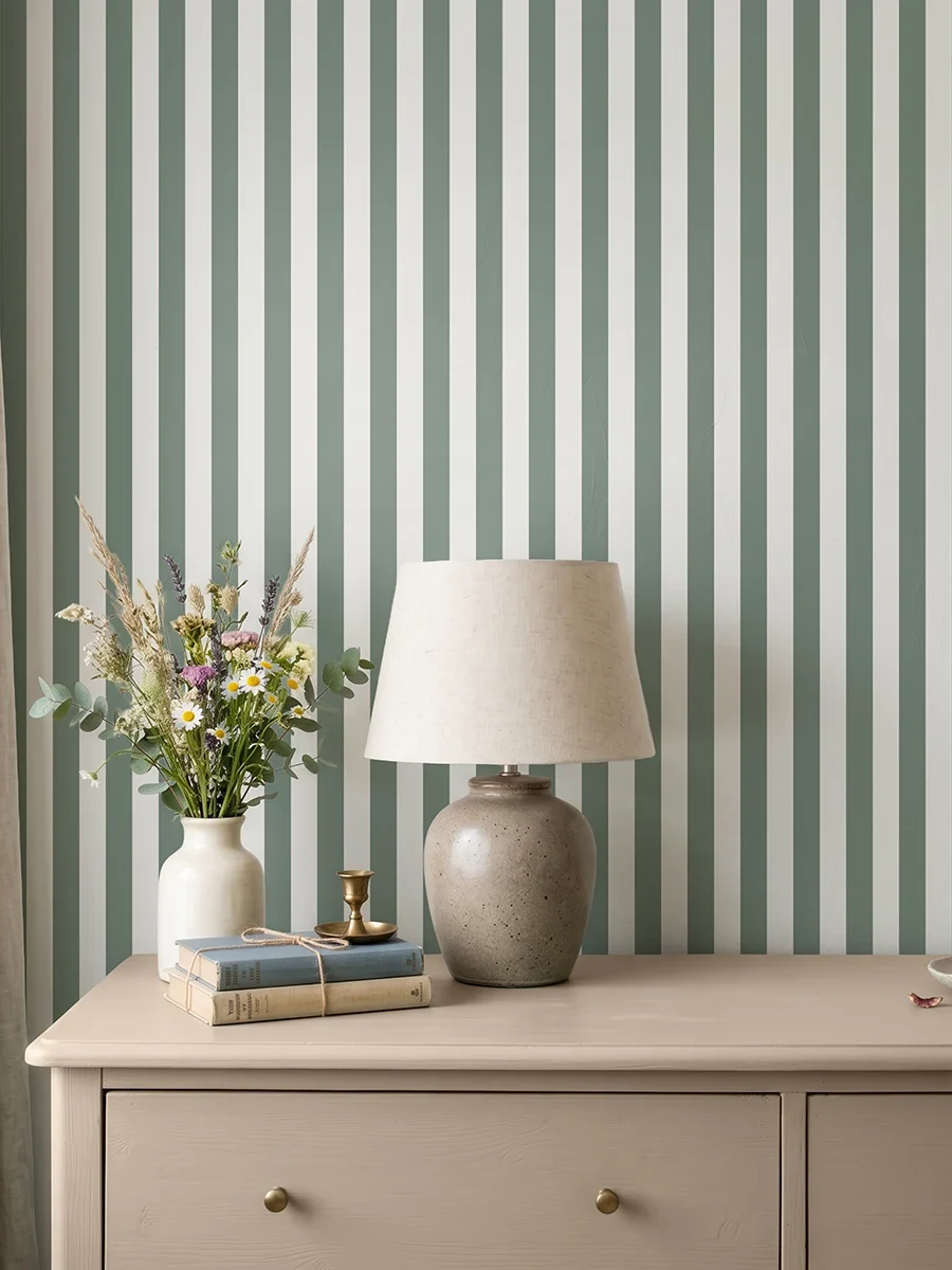 Modelo Della, papel pintado de rayas oliva y blanco lino, a escala pequeña en dormitorio con cómoda de madera clara, con una lámpara de sobremesa, algunos libros y un pequeño jarrón con flores.
