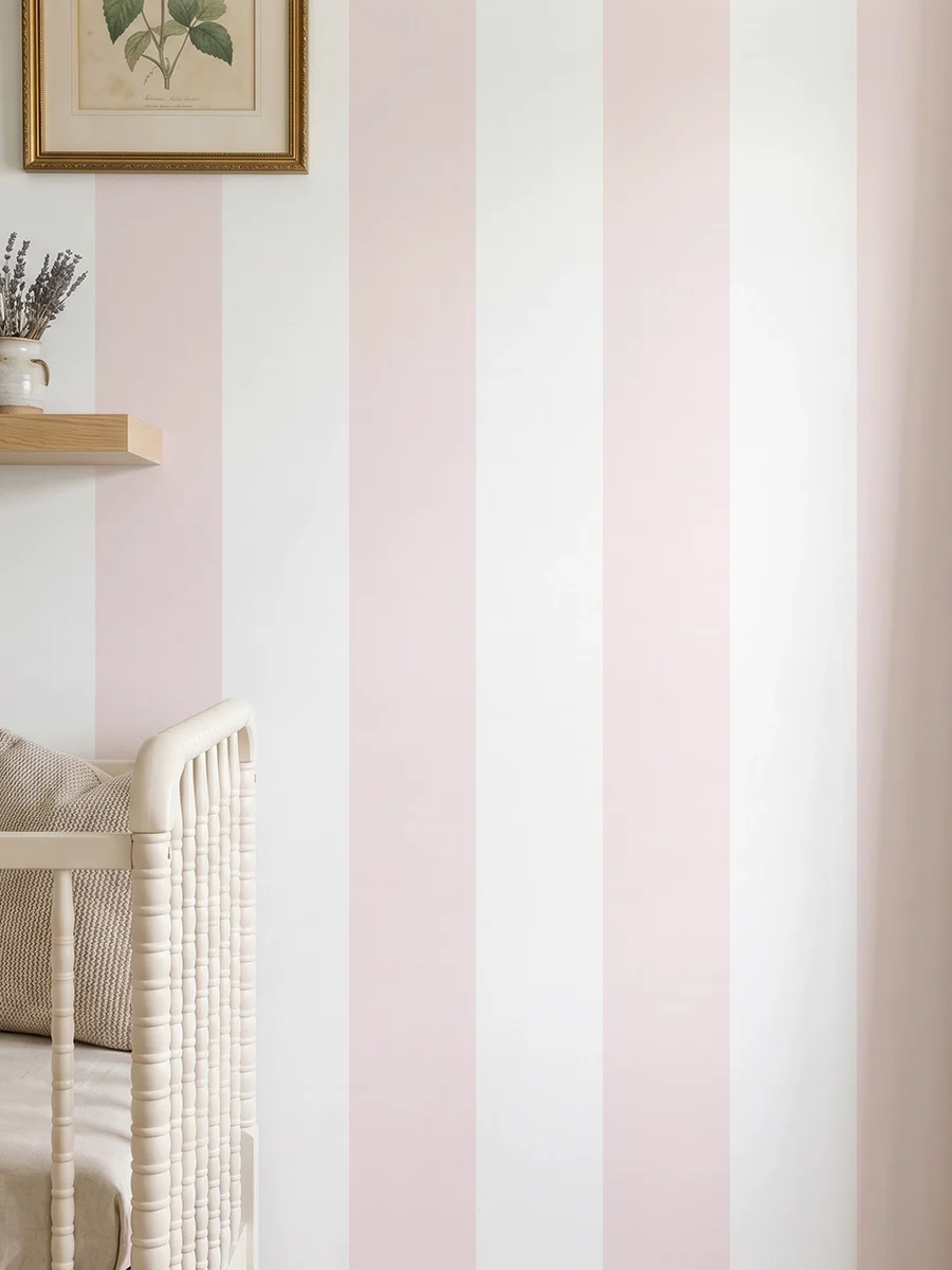 Modelo Della, papel pintado de rayas rosa nude y blanco, a escala maxi en dormitorio infantil con cuna de bebé y pared decorada con una pequeña estantería con elementos decorativos.