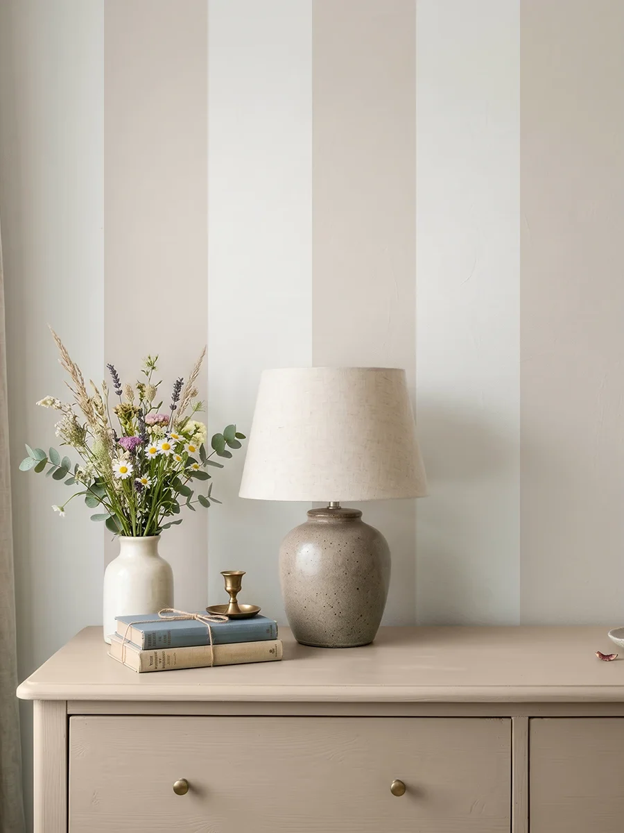Modelo Della, papel pintado de rayas beige y blanco lino, a escala maxi en dormitorio con cómoda de madera clara, con una lámpara de sobremesa, algunos libros y un pequeño jarrón con flores.
